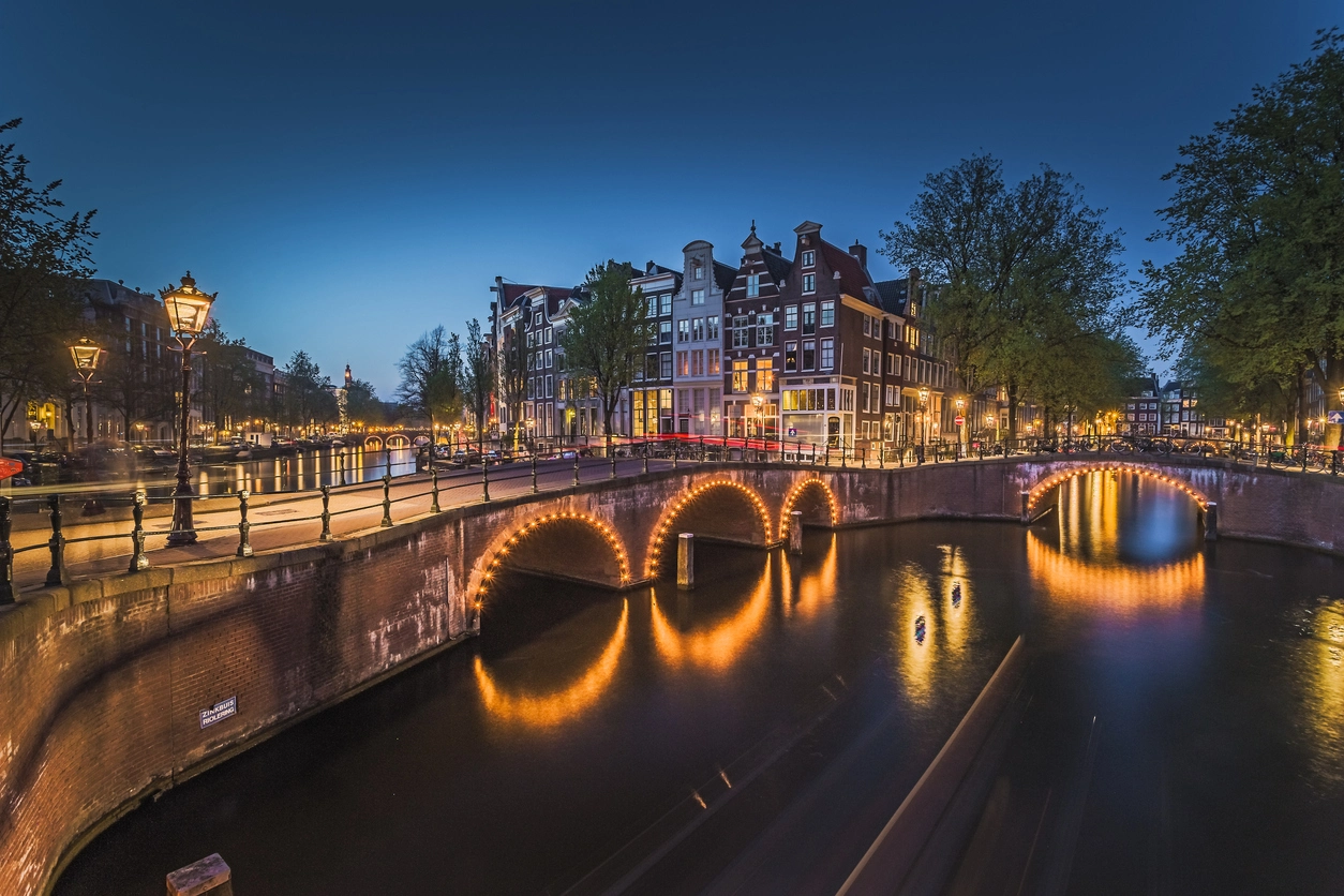 A twilight cityscape featuring illuminated bridges over a canal, flanked by historic buildings and streetlights, with a clear blue sky above.