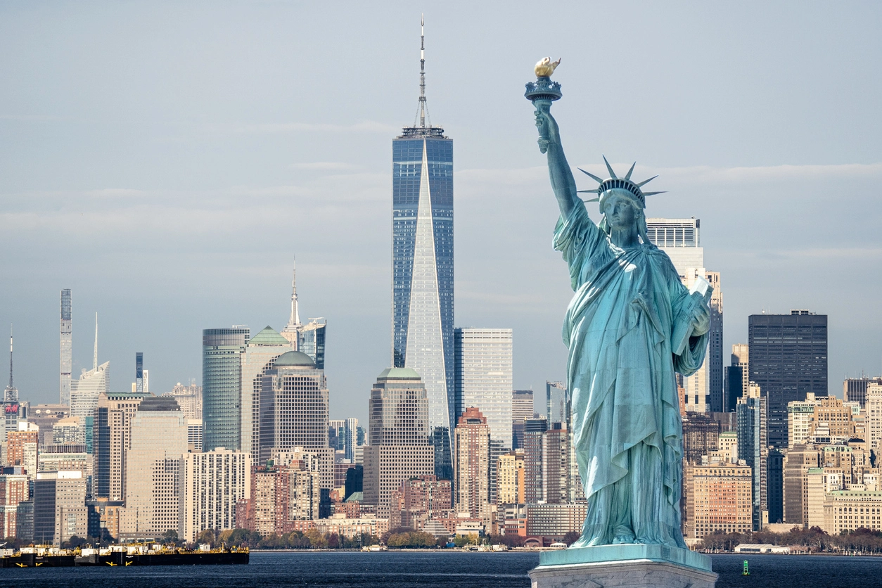 The Statue of Liberty stands in the foreground, with the New York City skyline and One World Trade Center in the background.