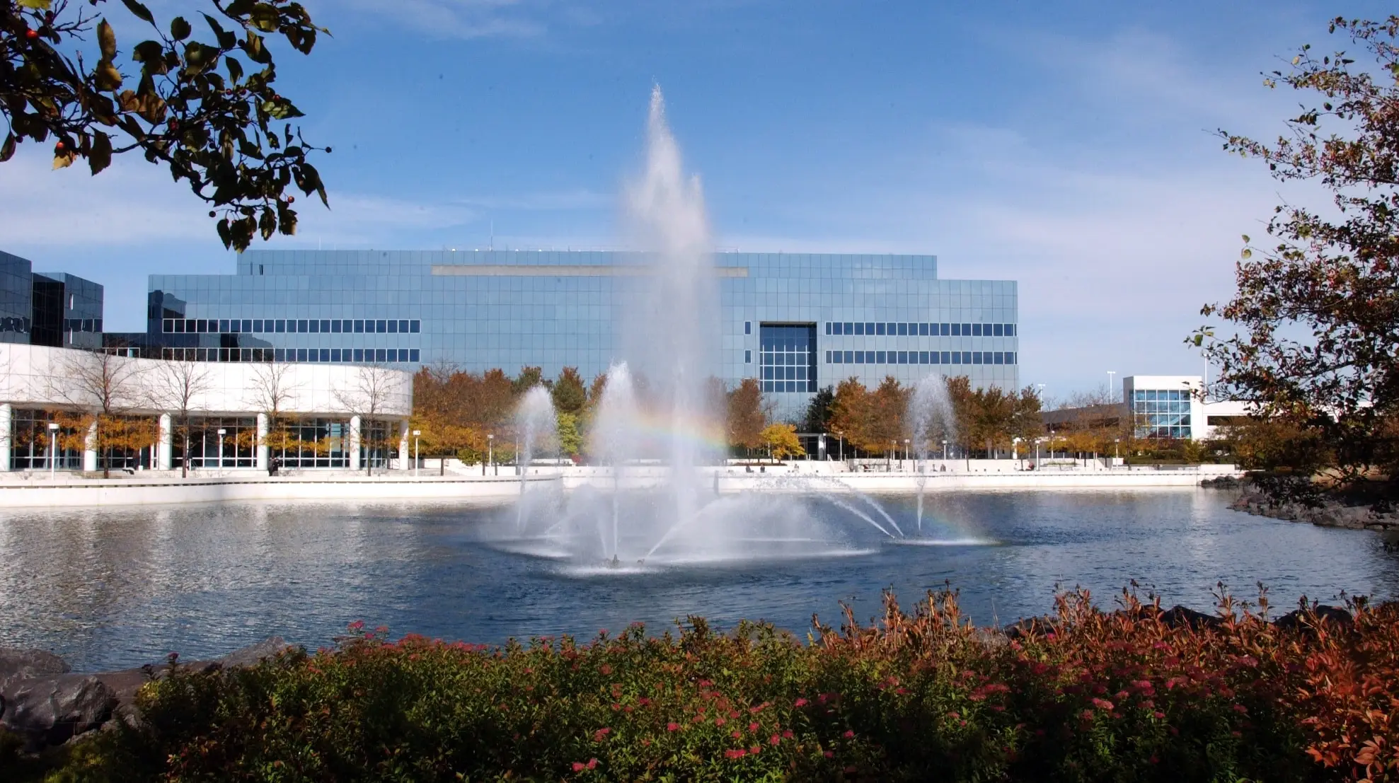 Sears Demolition featured image: A large fountain in a pond before a modern glass building, surrounded by autumn foliage under a clear blue sky, with a visible rainbow in the mist.